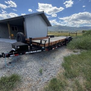 Flatbed trailer with wooden deck and spare tire parked on gravel near a metal shed in a rural yard with mountains in the distance.