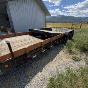 Long flatbed trailer with wooden side rails and a folded ramp, parked on gravel beside a metal shed in a rural field.