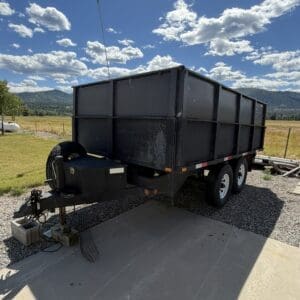 Black dump trailer with dual axles attached to a hitch on a gravel driveway, with a rural field and blue sky in the background.