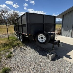 Black dump trailer with white wheels parked on gravel beside a metal shed under a blue sky.