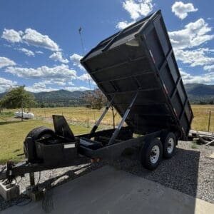 Dump trailer with raised bed supported by hydraulic pistons on a gravel lot, ready to unload in a rural field.