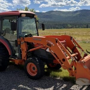 Orange Kubota tractor with a front-end loader and large bucket, parked on a gravel path with a rural field and forested hills in the background.