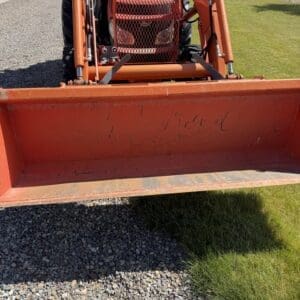 Orange front-end loader attachment mounted on a tractor, parked on gravel with green field in background, dirt in the bucket interior.
