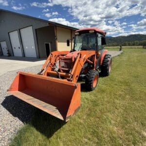 Orange front-loader tractor parked on a grassy field beside a metal building under puffy blue sky with scattered clouds.