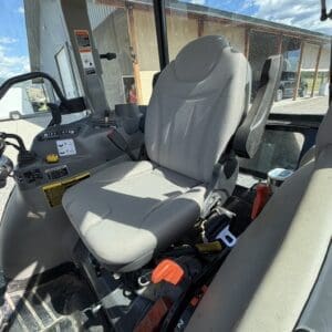 CAB interior of a tractor: front driver seat with grey upholstery, steering wheel, and control panel visible; outdoors a building in the background.