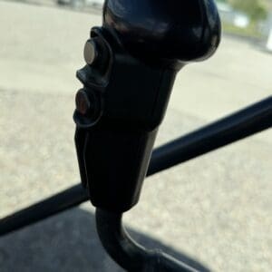 Black joystick-style control with two round buttons, mounted in a vehicle cab with an outdoor background blurred behind it