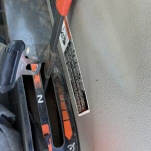 Close-up of an orange clamp/lever inside a vehicle, attached to a panel with a warning label and numeric indicator nearby (text on label unreadable).