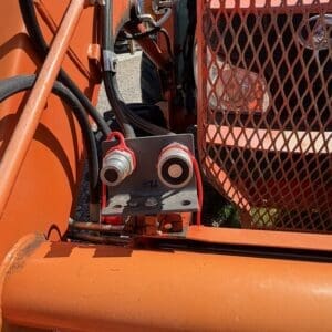 Close-up of hydraulic hoses and fittings on orange industrial equipment, with a small mounting plate and two round ports.