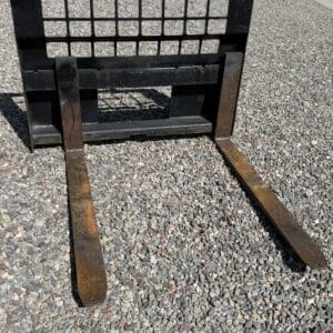 Metal forklift carriage with two long rusted forks resting on gravel outdoors.