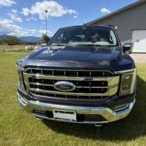 Front view of a dark blue Ford pickup with a chrome grille, parked on a grassy field near a metal barn.