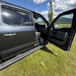 Dark pickup truck with both front and rear doors open, parked on a grassy field under a blue sky with mountains in the distance.