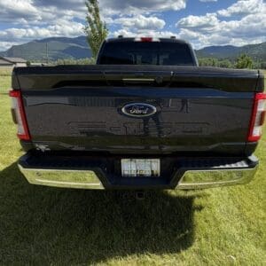 Rear view of a black Ford pickup truck parked on a grassy field with mountains under a blue, cloudy sky in the background.