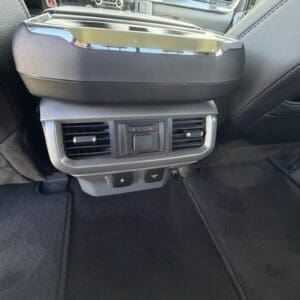 Rear-seat area of a car showing the back of the center console: two air vents, a small control panel, and a folded armrest above with carpeted floor beneath.
