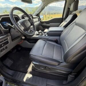 Interior of a black pickup truck cabin: driver and front passenger seats, steering wheel, center console, and dashboard bathed in daylight with a mountain landscape visible through the windows.
