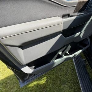 Open car door interior showing a gray panel, armrest, small storage pocket, and window controls; grass outside the vehicle.