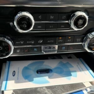 Center console with circular volume and tuning knobs and row of climate control buttons above Ford owner's manuals in the glove compartment.