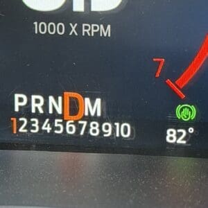 Close-up of a car dashboard showing gear indicator PRNDM with the D highlighted in orange, the numeric gear positions 1–0 beneath, and speedometer/tachometer text reading 1000 X RPM with 82° nearby and a green function icon nearby.