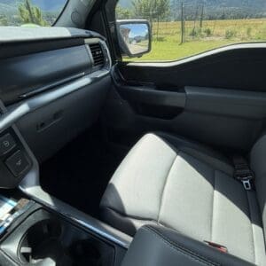 View from the front passenger seat of a gray-trimmed vehicle, showing the center console and cup holders with a countryside scene outside (fence, trees, hills).