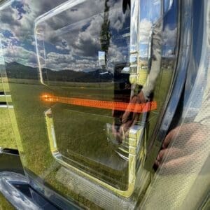Close-up of a truck headlight with an amber turn signal, chrome trim, reflecting a cloudy sky and green field in the chrome surface.