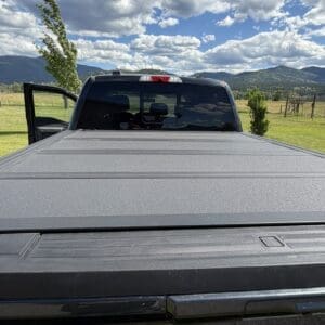 Rear view of a black pickup with a closed tonneau cover, driver’s door open, in a grassy field with mountains and a cloudy sky behind it.”—informative image