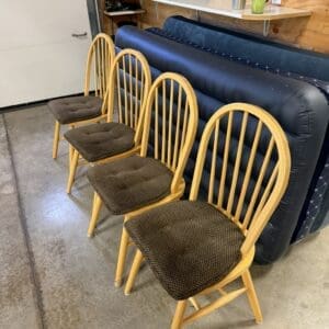 Four wooden chairs with brown cushioned seats lined up in a garage workshop, with stacked blue air mattresses behind them.