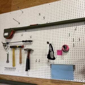 Pegboard wall in a workshop with organized hand tools: hammers, mallets, a wrench, tape measure pouch, and safety goggles, plus a blue folder.