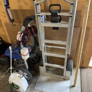 Aluminum hand truck with a curved top, two wheels, and a black strap resting against a plywood wall in a cluttered garage.