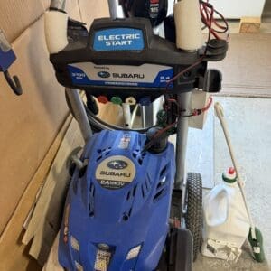 Blue Subaru EA190V snow blower with an electric-start panel labeled ELECTRIC START, gas can nearby, and various colored control knobs in a cluttered garage.