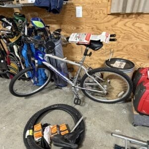 A cluttered garage with a silver mountain bike leaning against a wooden wall, surrounded by gear including golf bags, a spare tire with orange-wrapped parts, and a red portable machine on the right.