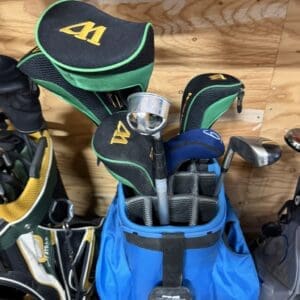 Blue golf bag with grey dividers holding several clubs, green-black headcovers with yellow logos, and a metal putter ferrule, leaning against a wooden wall.