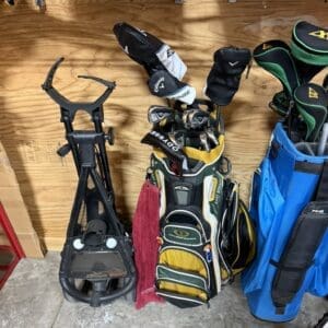 Black golf push cart on the left and several golf bags with clubs leaning against a wooden wall in a storage area, with a red towel nearby and a blue bag to the right.