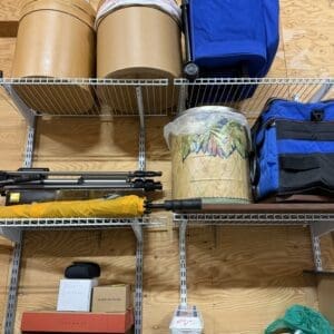 Cluttered storage shelving with large brown cylinders, blue bags, tripods, and boxes on wooden shelves.