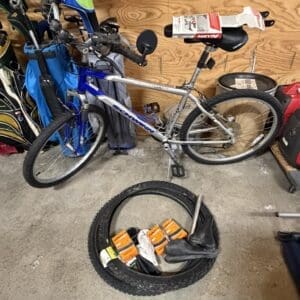 A cluttered garage with a silver bicycle parked against a pegboard wall and a tire sitting on the concrete floor containing patch kits and gloves inside it.