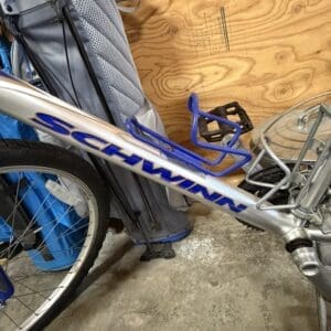 Left side view of a Schwinn bicycle frame with blue bottle cages, parked in a garage beside a gray suitcase against a plywood wall.