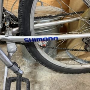 Close-up of a silver Shimano mountain bike frame with the rear wheel, chain, and pedal visible in a garage setting with a metal bucket in the background.
