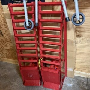 Two red metal rolling stair trolleys standing upright in a workshop, leaning against plywood and cardboard boxes.