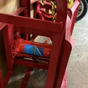 Red hydraulic bottle jack stored in a metal frame rack in a workshop, with yellow-and-black caution tape nearby.
