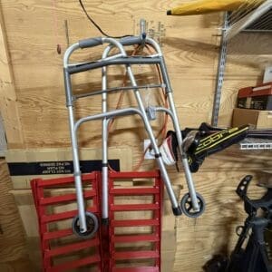 Metal rolling walker resting on two red metal stair ramps leaning against a plywood wall in a cluttered workshop/garage setting.