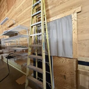 Tall yellow extension ladder with a wheeled base leaning against a wooden workshop wall near metal shelves.