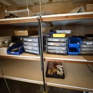 Tall black telescoping pole with a yellow foot pad standing upright in front of a workbench with plastic storage drawers and bins in a workshop.