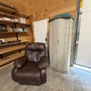 Brown leather recliner chair in a wood-paneled garage with shelves storing cans and tools, next to a large gray storage shed and an open garage door.