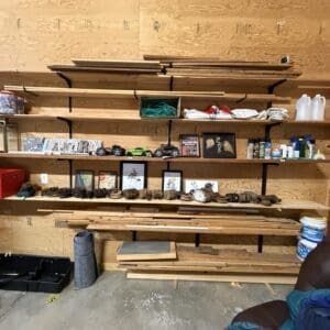 Woodworking shop shelves holding lumber, containers, and small framed items on a pegboard wall.