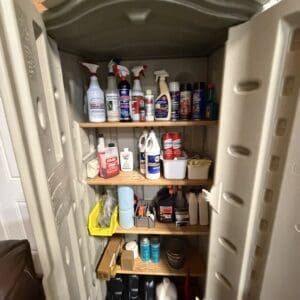 Open storage cabinet filled with cleaning products on three wooden shelves: spray bottles, detergents, and household cleaners in a cluttered arrangement.
