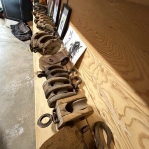 Row of rusty vintage pulleys and iron blocks lined along a wooden workbench, with framed drawings leaning against the wall.