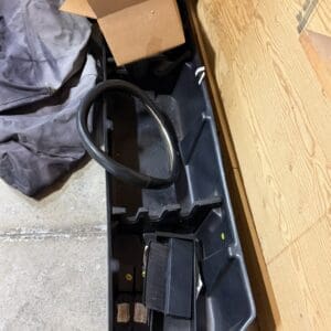 Open plastic tote on a concrete floor filled with a coiled black hose, a cardboard box, and miscellaneous parts.