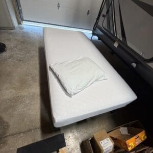 White mattress on a frame in a garage next to a travel trailer, with a folded white sheet on top and boxes on the concrete floor