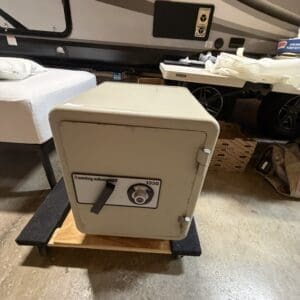 Heavy gray safe with lever and combination dial on a wooden pallet in a garage beside an RV trailer window and mattress nearby.