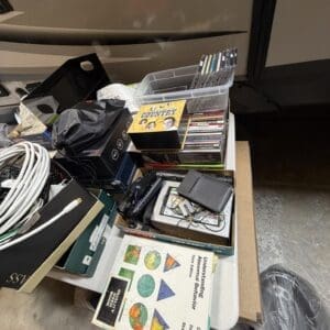 A cluttered collection of cables, boxes, DVDs, and books piled in and around a plastic bin on a table edge.