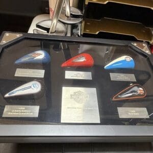 Six colored Harley‑Davidson gas-tank models in a glass display case, with a central plaque and small labels beneath each item describing the colors.