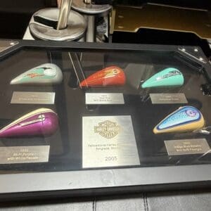 Display case with six colorful Harley-Davidson gas tank replicas mounted on a glass surface, each with metal plaques, and a central plaque for 2005.